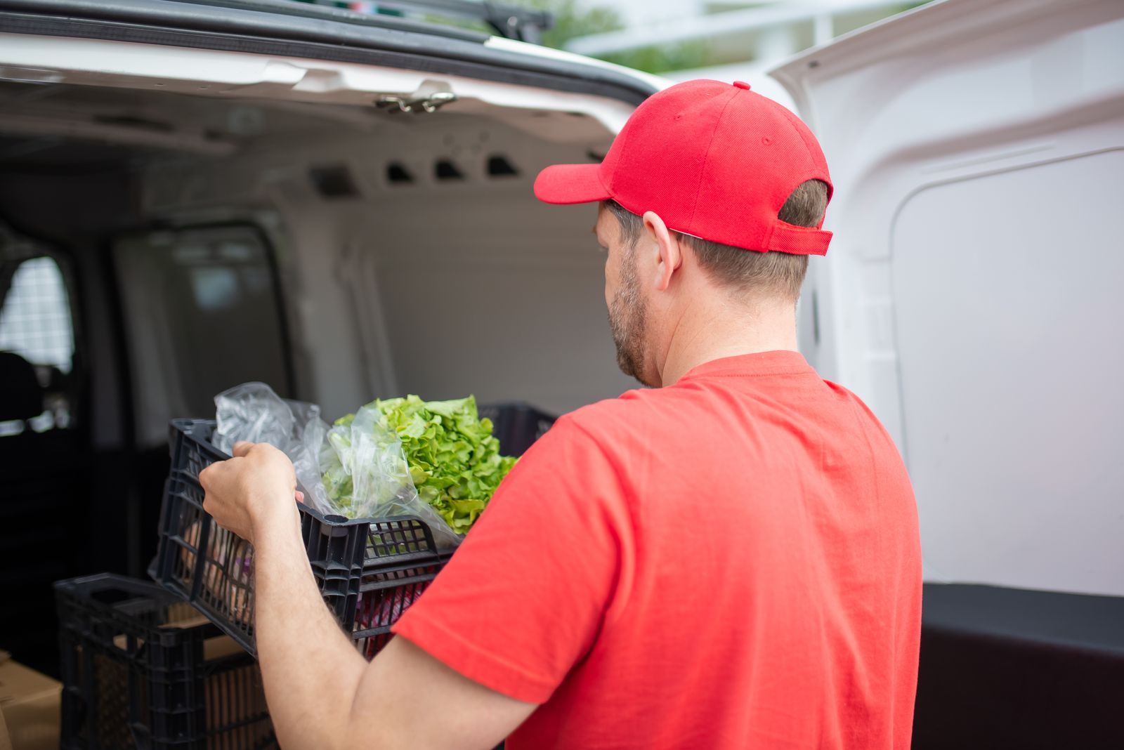 A delivery worker, seen from behind, lifting a crate of vegetables from a van